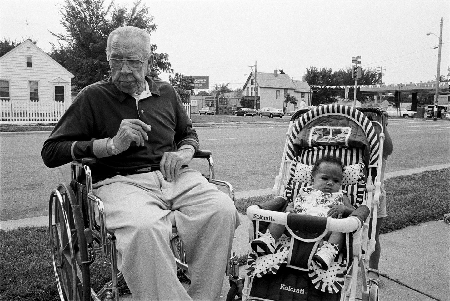 An old man in a wheelchair sits next to a baby in a stroller