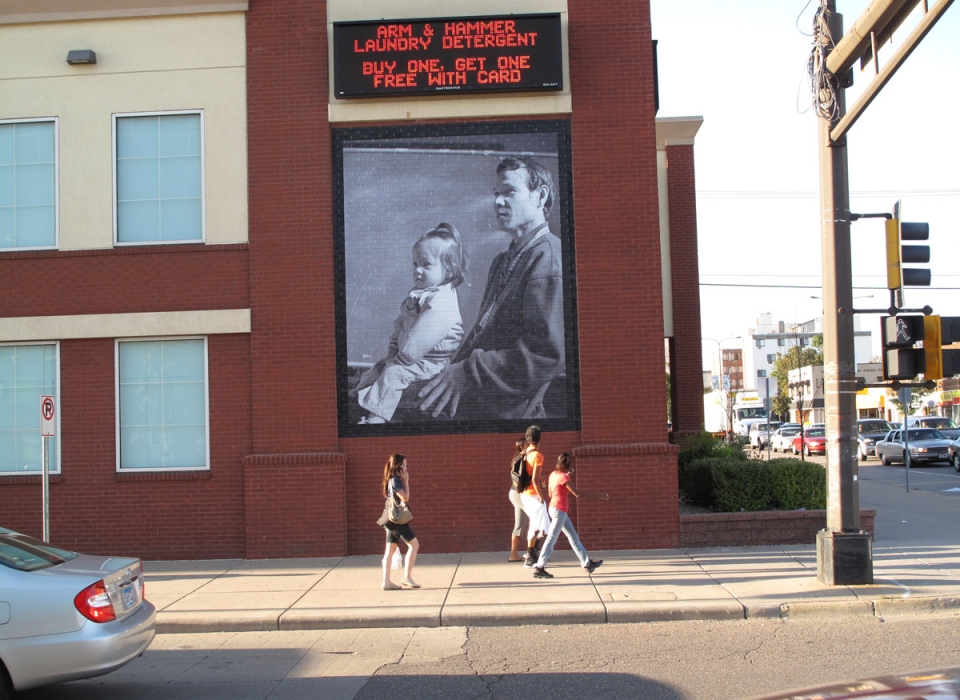 People walking down the sidewalk look at a large photo on the side of a building of a man with a child on his lap