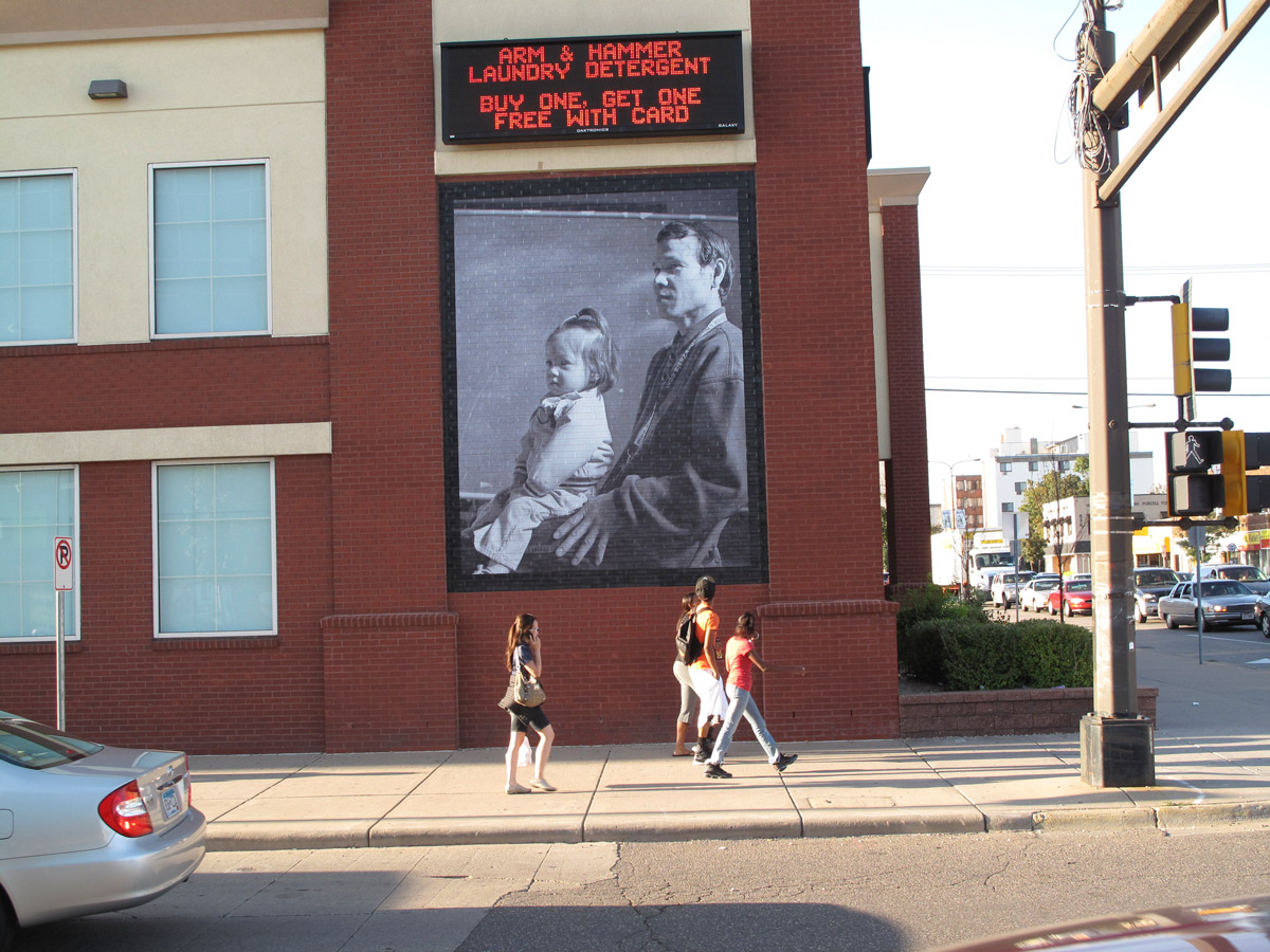 People walking down the sidewalk look at a large photo on the side of a building of a man with a child on his lap