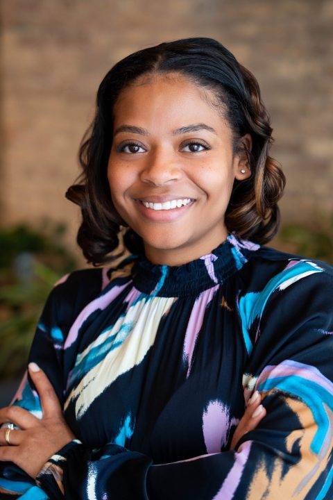 A headshot style photograph of a young woman in a colorful shirt, standing against a brick background.