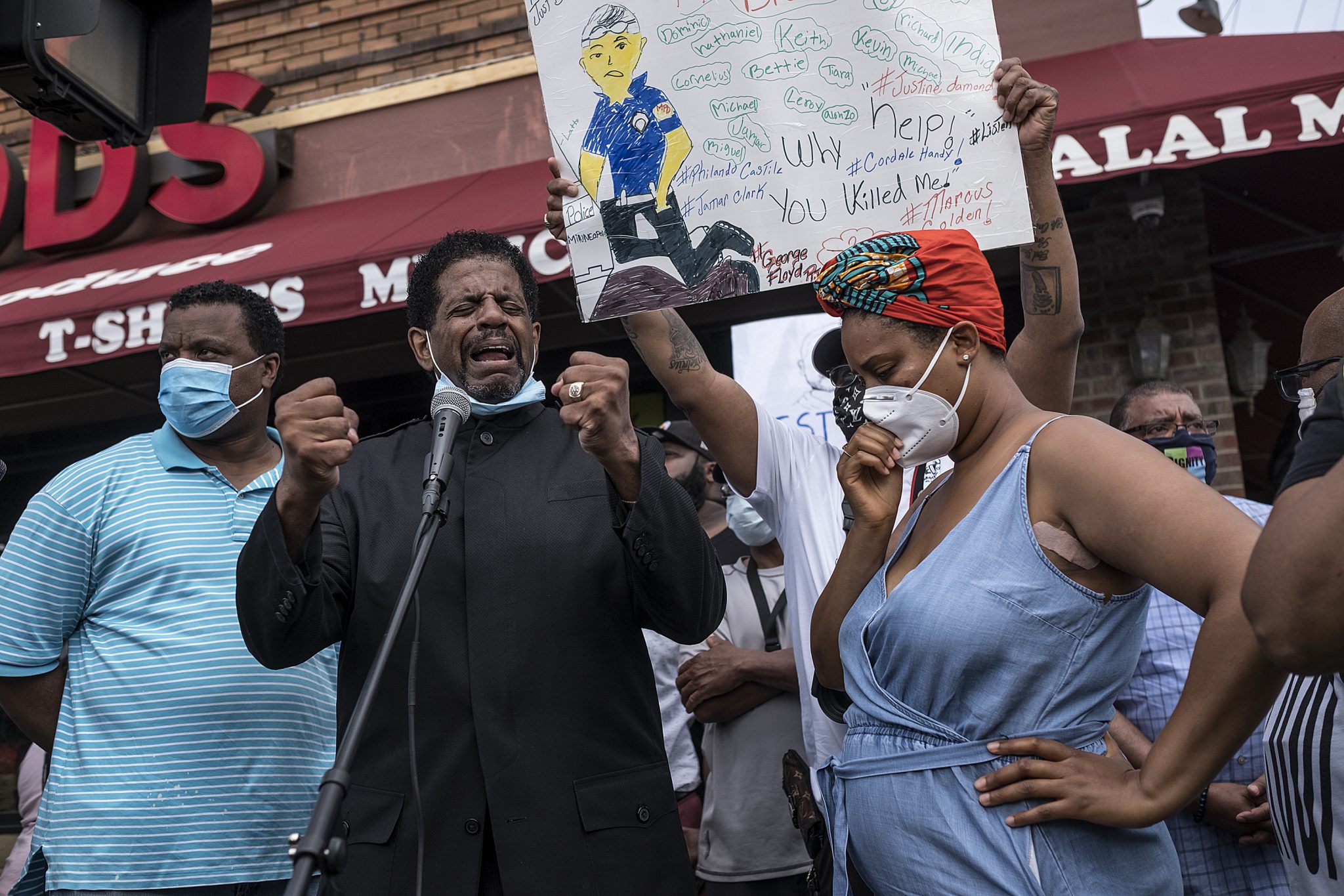 A group of people stand together, one holding a sign with handwritten messages and a drawing of a person. In the background, a storefront displays the words "T-SHIRTS" and "HALAL MEAT."