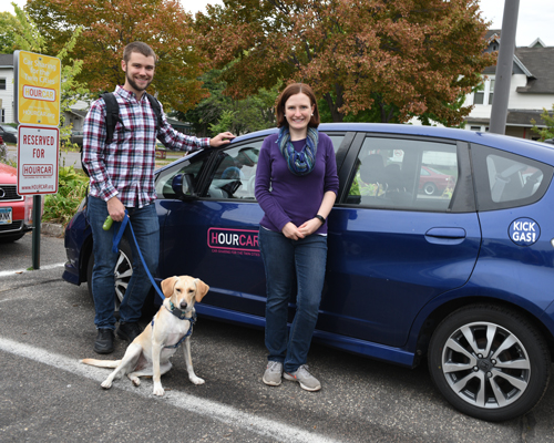 A man, and a woman stand in front of and electrical car, the car says Hourcar on the side