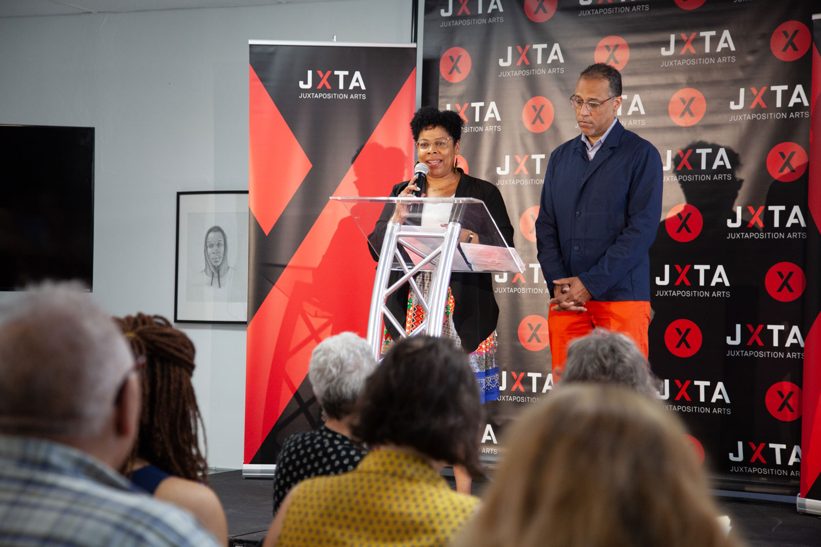 Two people stand at a podium and talk to a crowd.