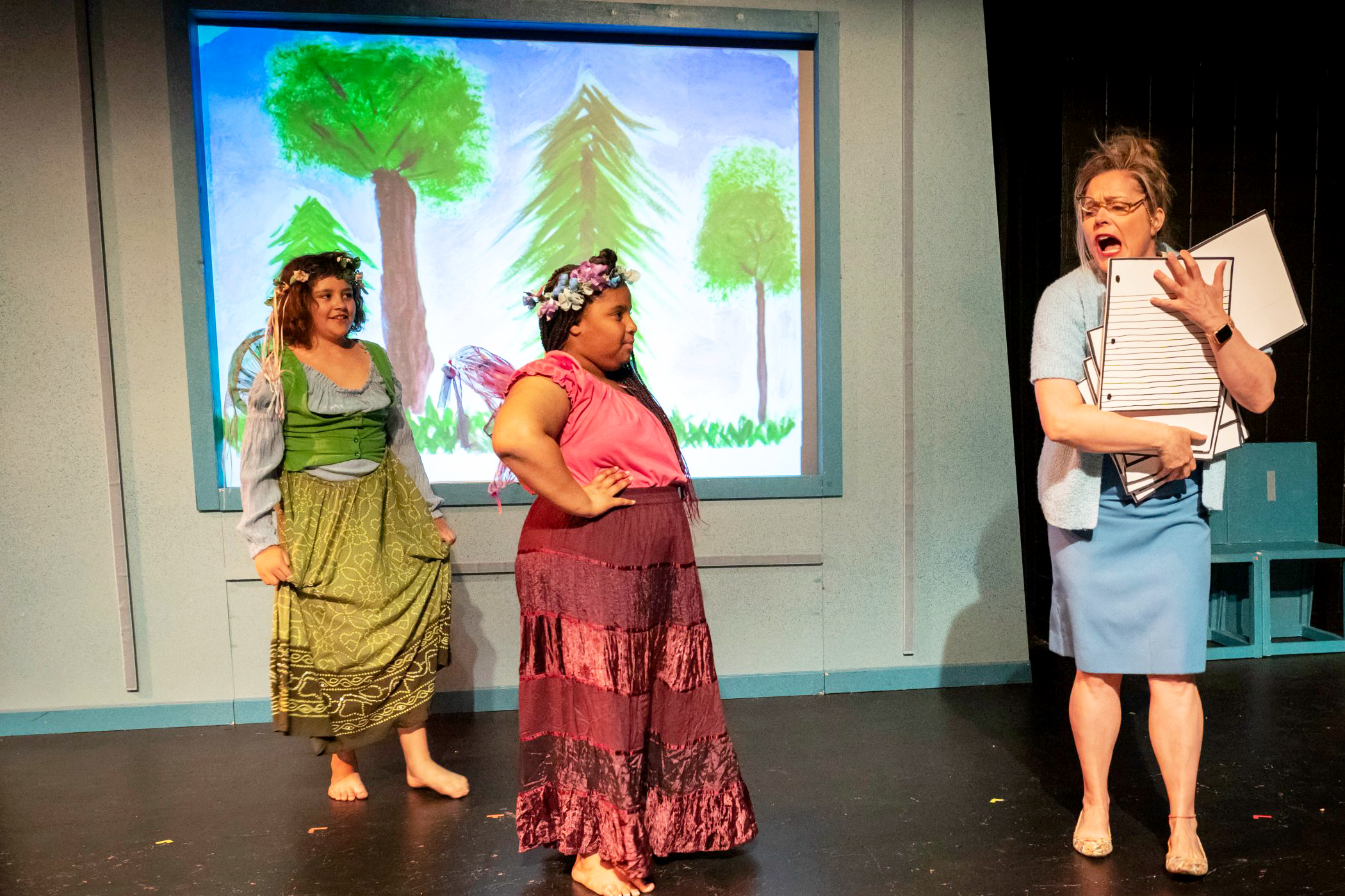 Two youth performers and one adult perform on a stage. A child's painting of trees is in the background. The adult performer appears to be playing the role of a teacher.