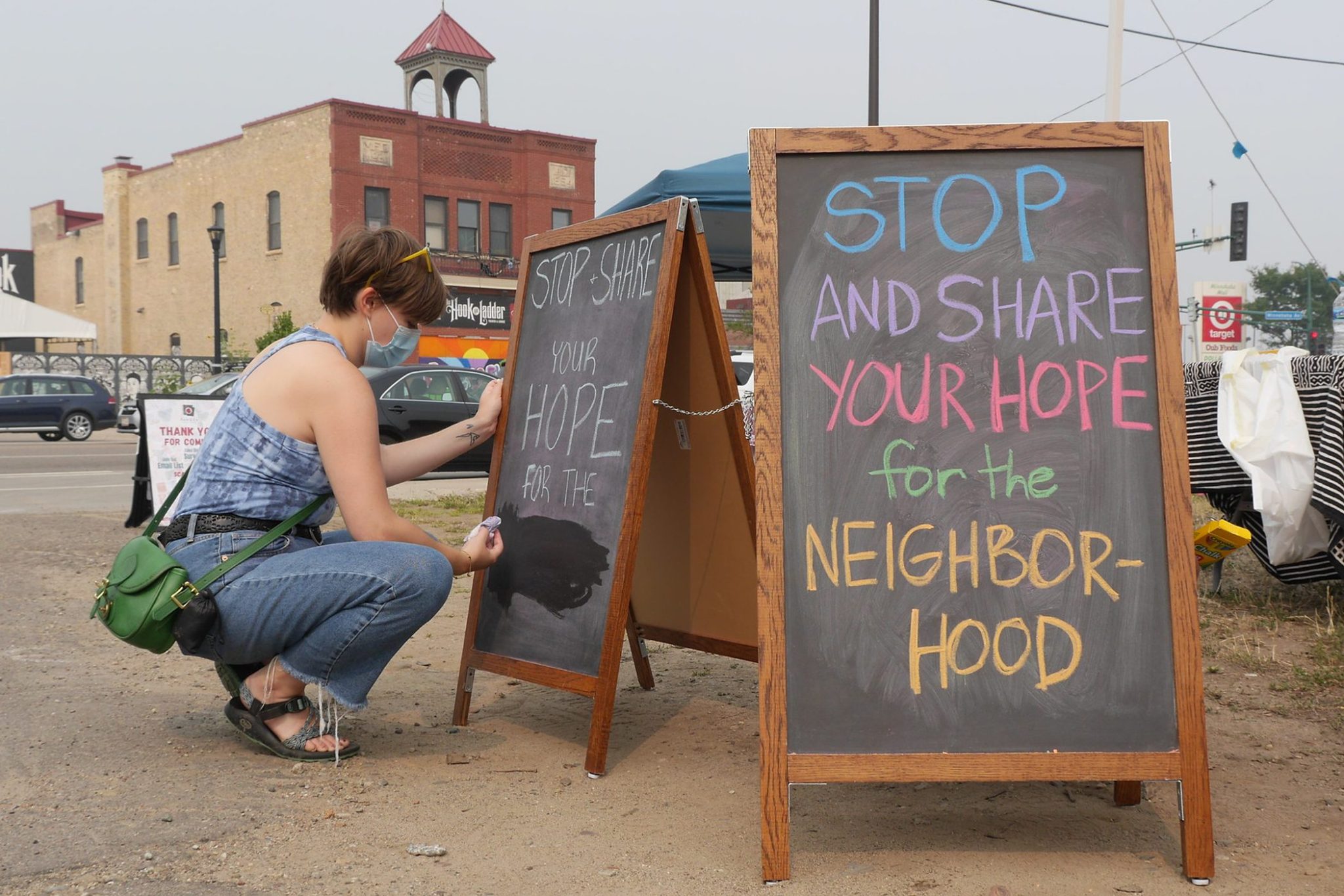 A person crouches and writes on a chalkboard with a large brick building in the background. A chalkboard in the foreground reads "Stop and share your hope for the neighborhood"