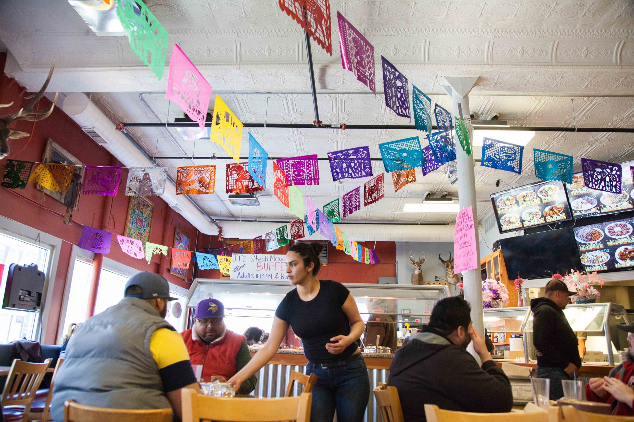 Kabalyeeri ayaa miiska ka soo saarta Plaza Del Sol. a waitress clearing a table, colorful flags hang from the ceiling.