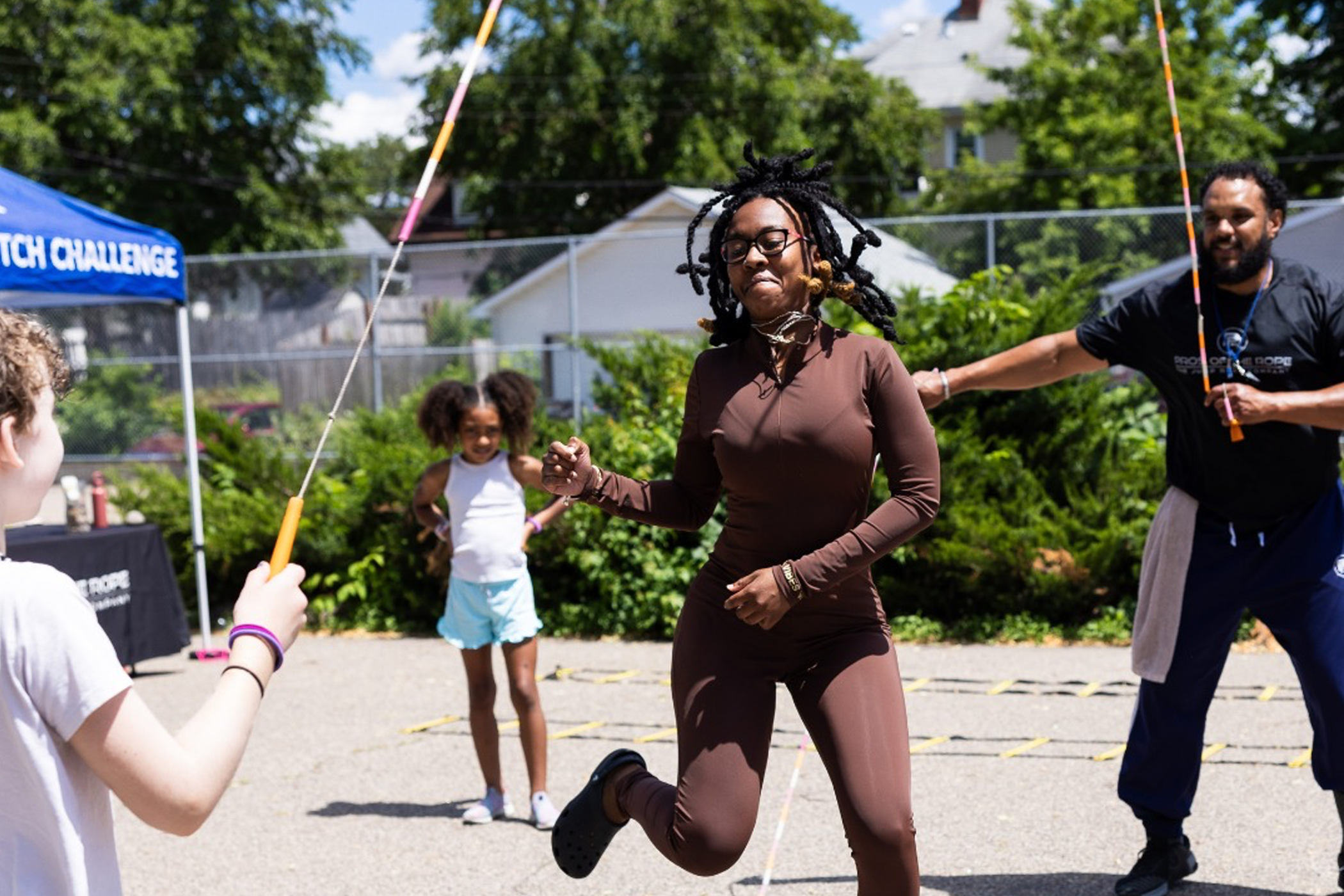 A group of people participate in a double dutch jump rope activity outdoors. One person jumps in the middle while two others turn the ropes, and another stands nearby watching. The background features greenery, a fence, and part of a building under sunny skies.