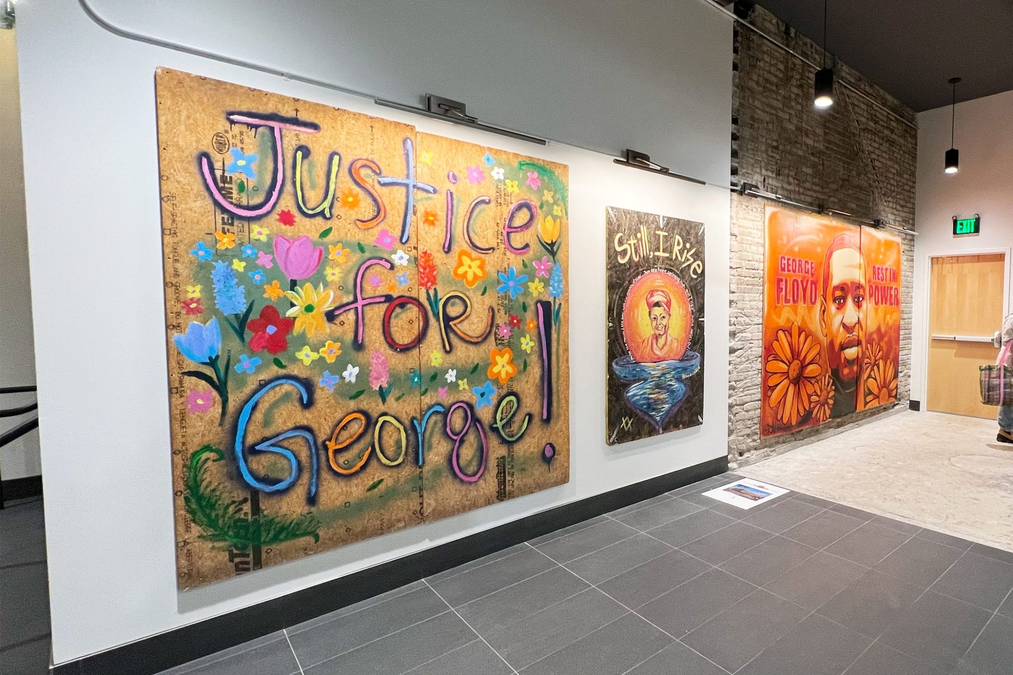 Three pieces of art hang on a gallery wall. Left to right: A floral graffiti-style piece on plywood that says "Justice for George!"; a painting of a Black woman with hand-painted words that read "Still, I Rise"; an orange and red painting of George Floyd that says 'George Floyd Rest in Power".