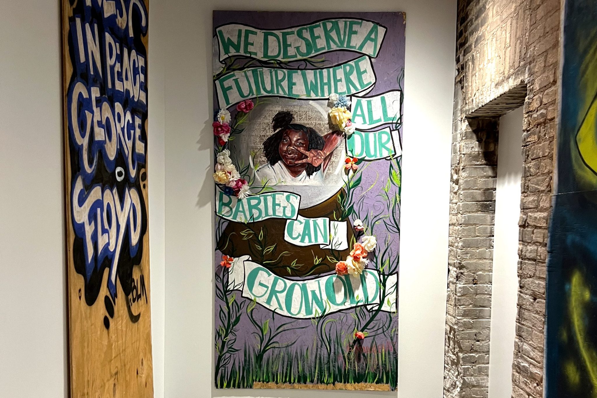 Two pieces of art hang on a wall. The piece on the left features graffiti-style text that reads "Rest in Pease George Floyd". The focal point of the photo is a piece of art that features a painting of a young Black woman making a peace sign with her hand, and hand-painted text that reads, "We Deserve a Future Where All Our Babies Can Grow Old"