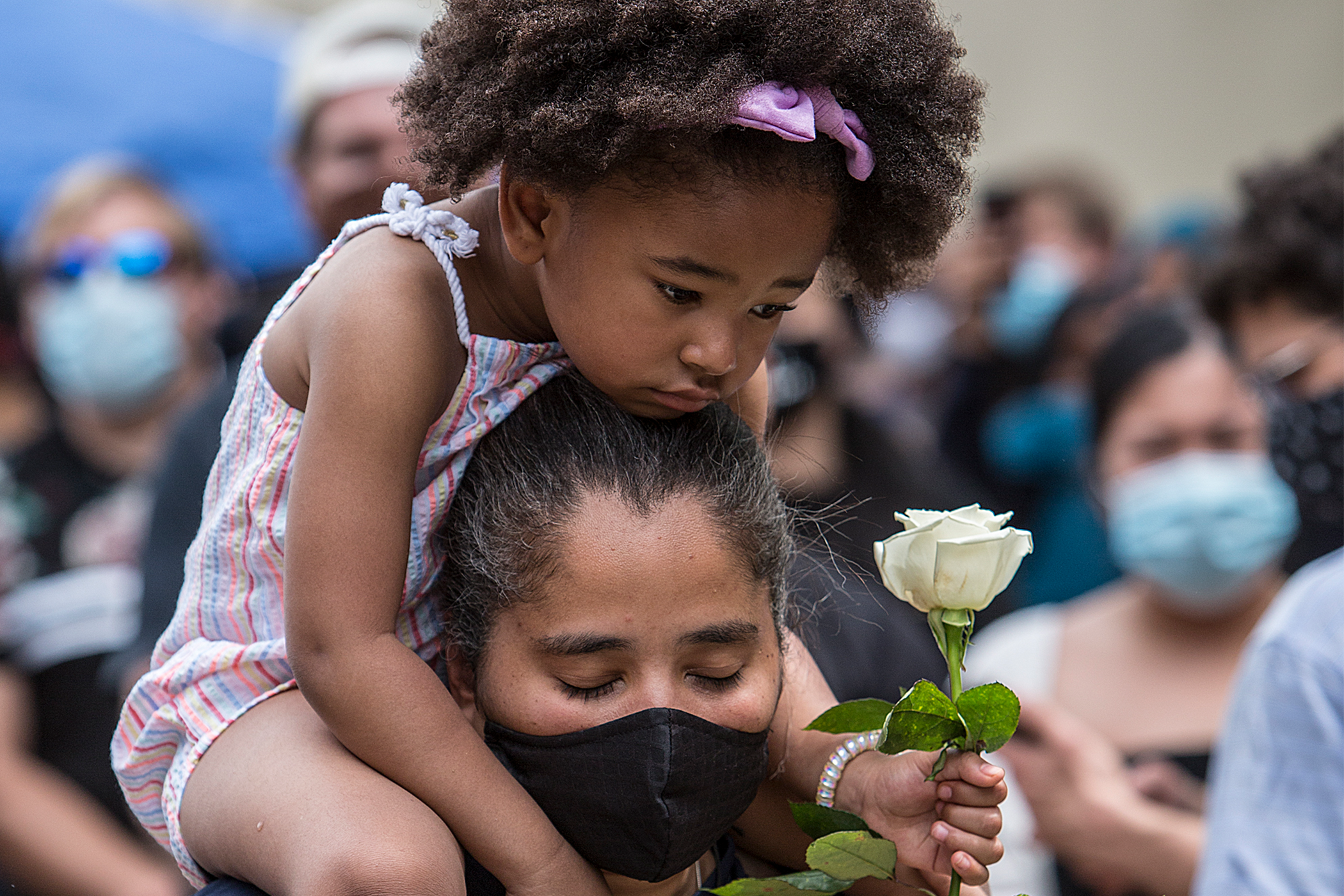 A child sits on the shoulders of a woman in a face mask, holding a rose with downturned eyes