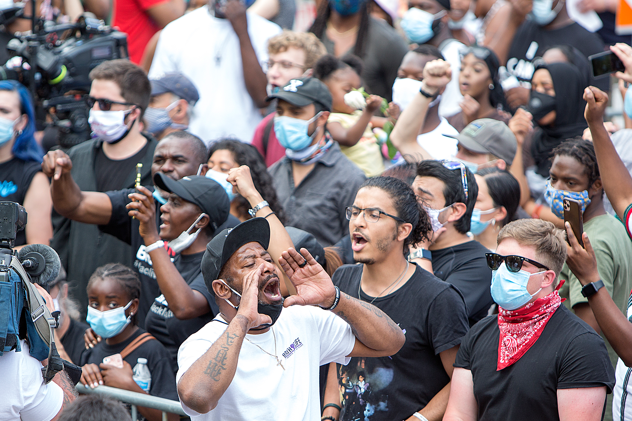 A large group of people gathered with fists raised in the air; some are wearing masks, and a person with a camera is visible on the left