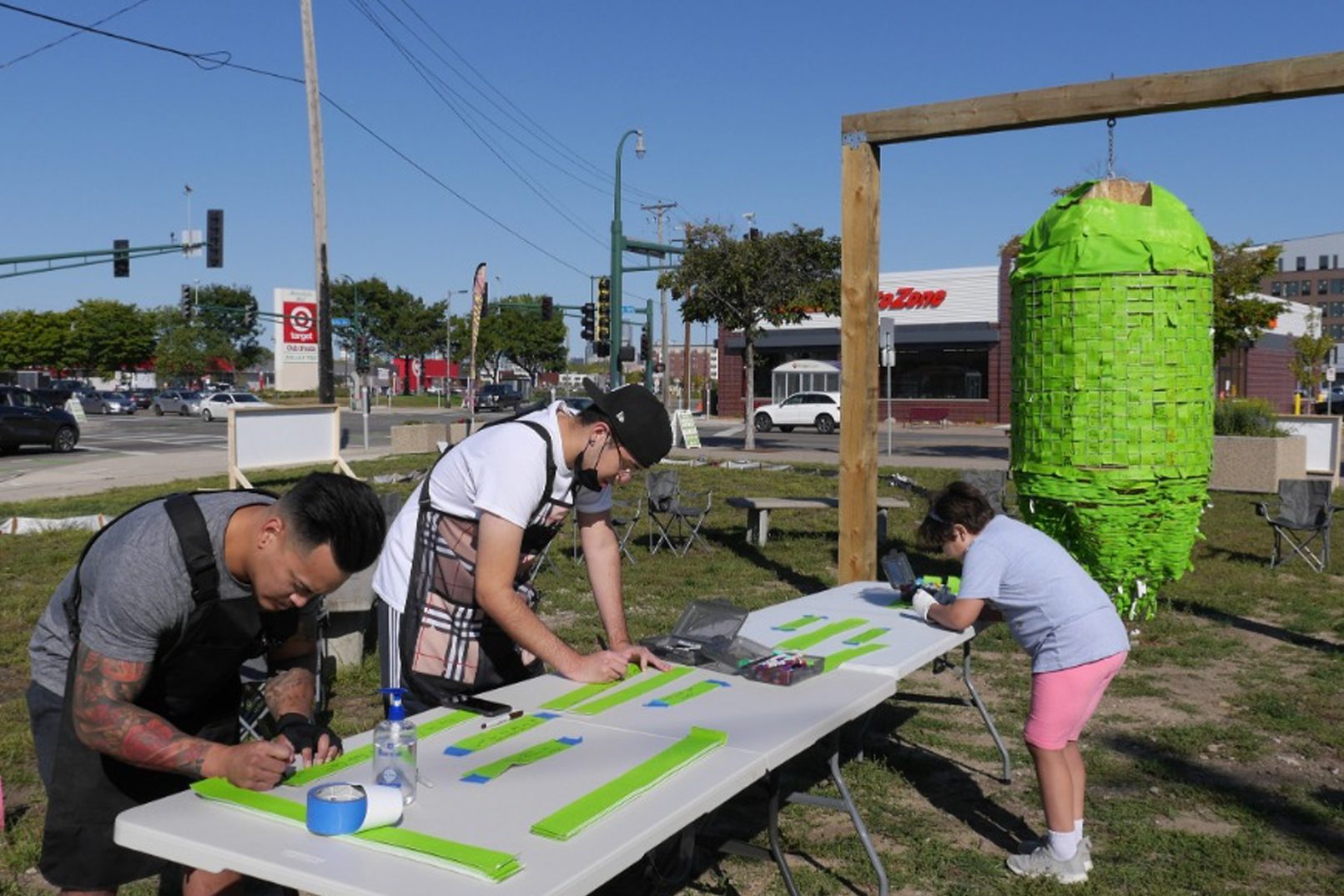 Three people write on strips of green paper at a folding table outdoors. To the right of the photo, a large sculpture comprised of the green strips of paper hangs from wooden beams.