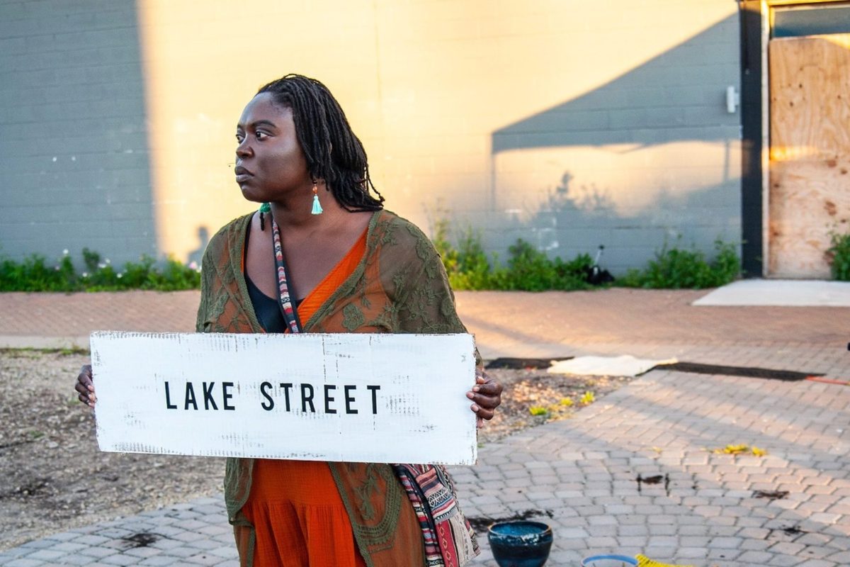 A Black woman stands outdoors holding a hand-painted sign that says "LAKE STREET"