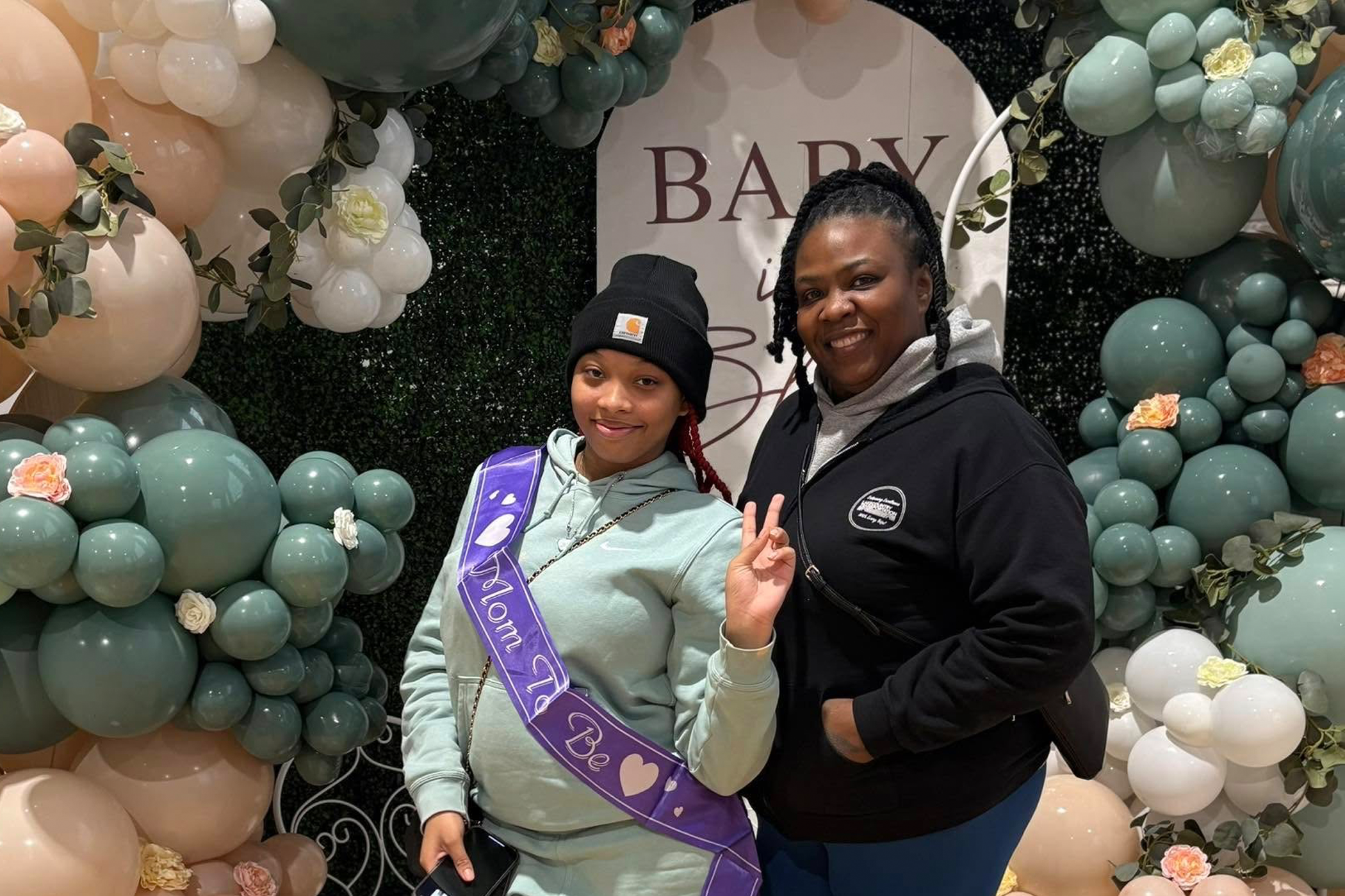 Two Black women stand in front of a partially visible sign that says "Baby," surrounded by balloons and flowers. The woman on the left wears a purple sash that says 'Mom to be."