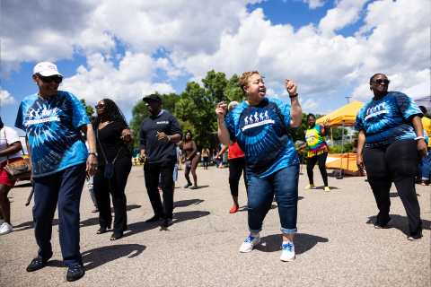 A group of people dance in a line on the street. They are wearing matching blue tie-dye shirts.