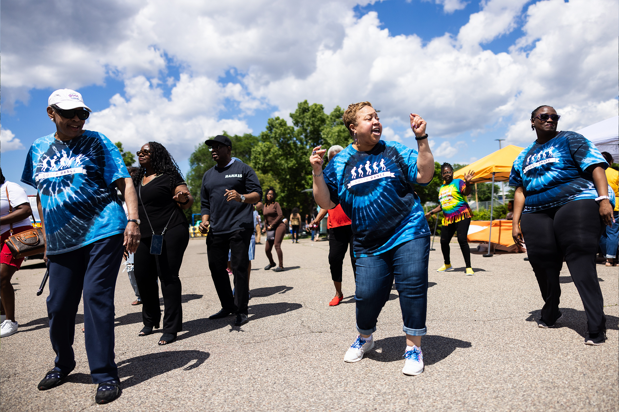 A group of people dance in a line on the street. They are wearing matching blue tie-dye shirts.
