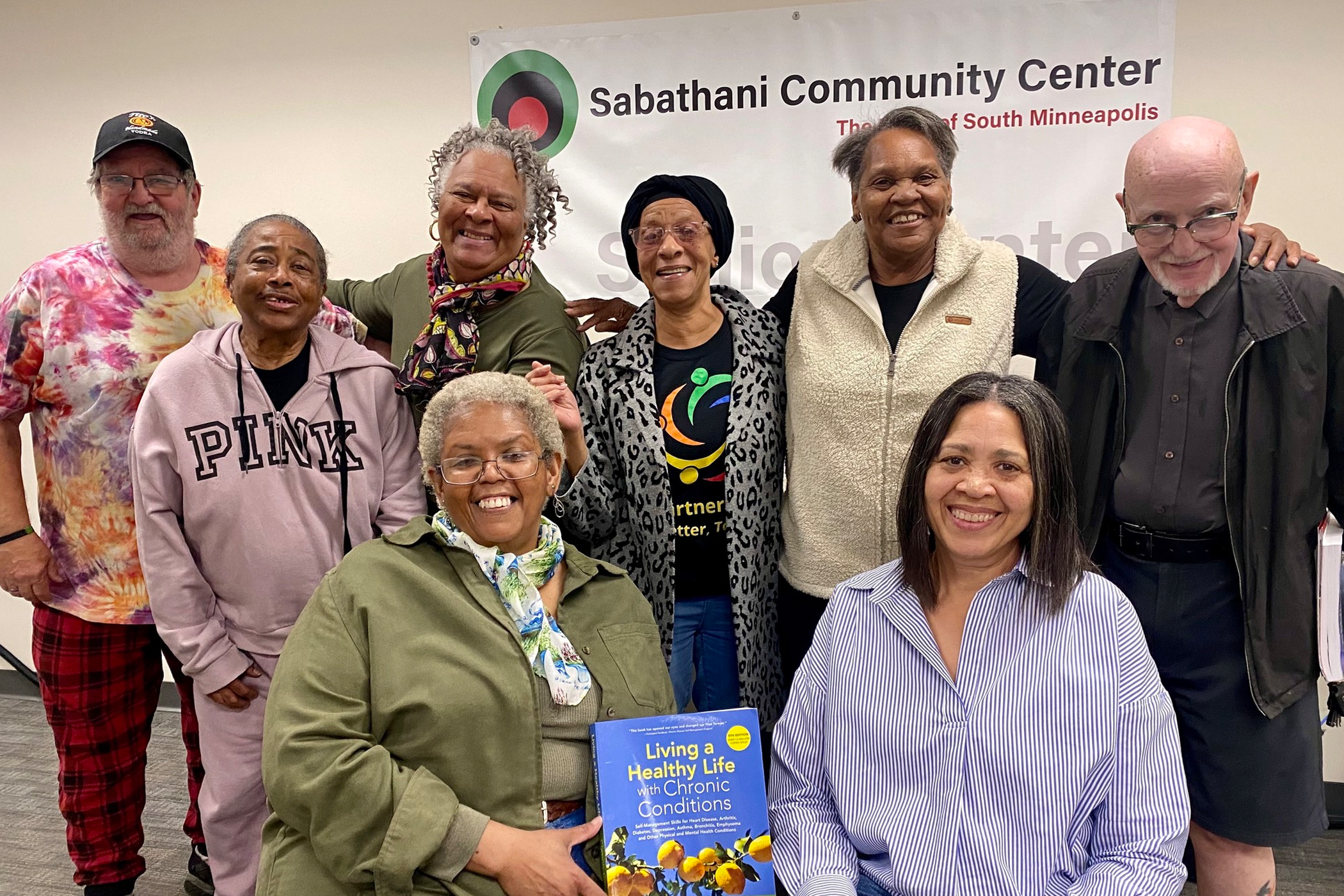 8 older adults pose in front of a sign that reads Sabathani Community Center. One person in front holds a book titled "Living a Healthy Life with Chronic Conditions."