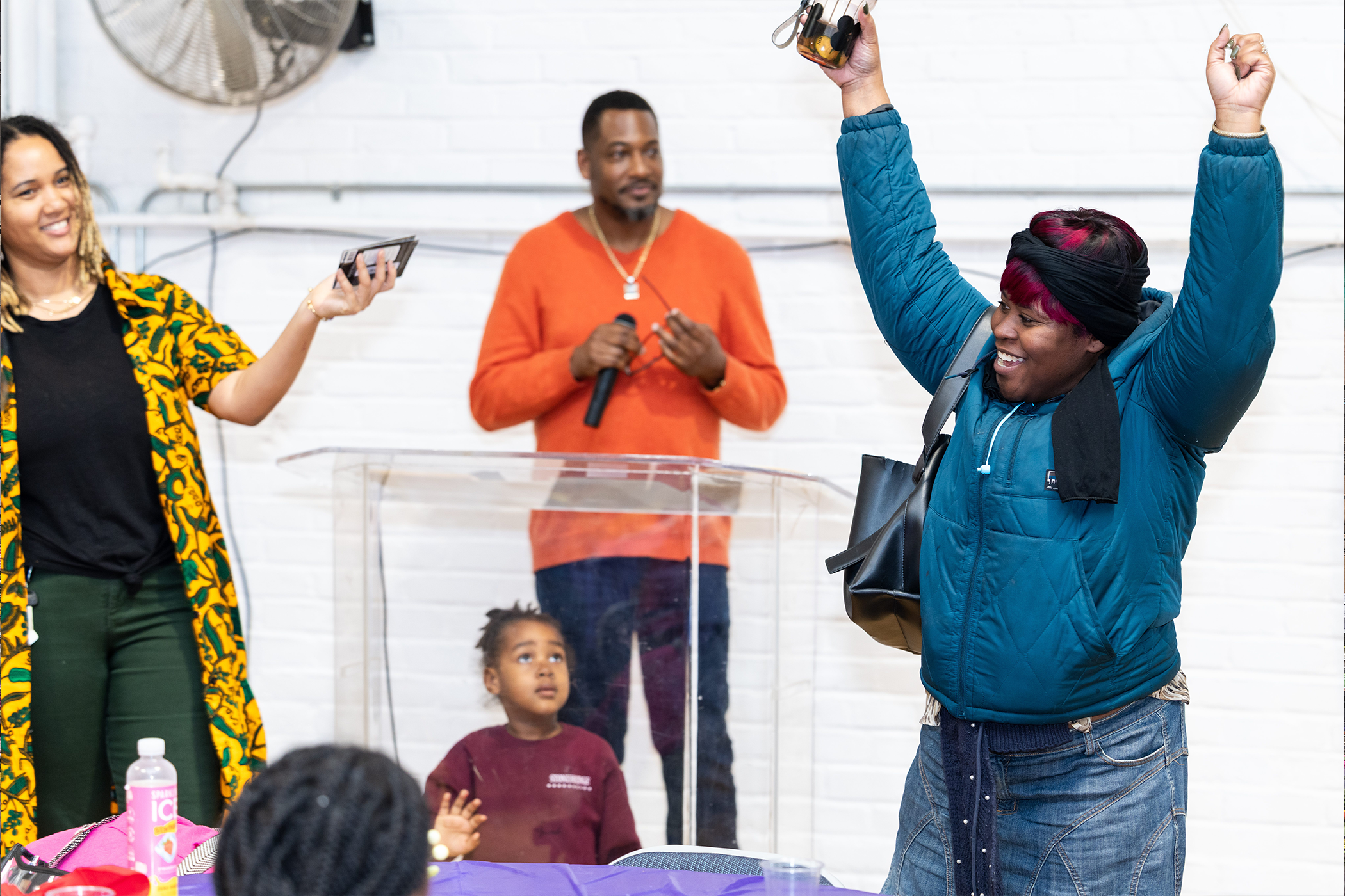 A group of people in an indoor setting, with one person speaking at a clear podium holding a microphone. Another person holds up a phone and an object, possibly recording, while a third raises both arms in the air. Two children are also present—one seated at a table and another standing near the podium. The background includes a white brick wall, electrical wiring, and an industrial fan.