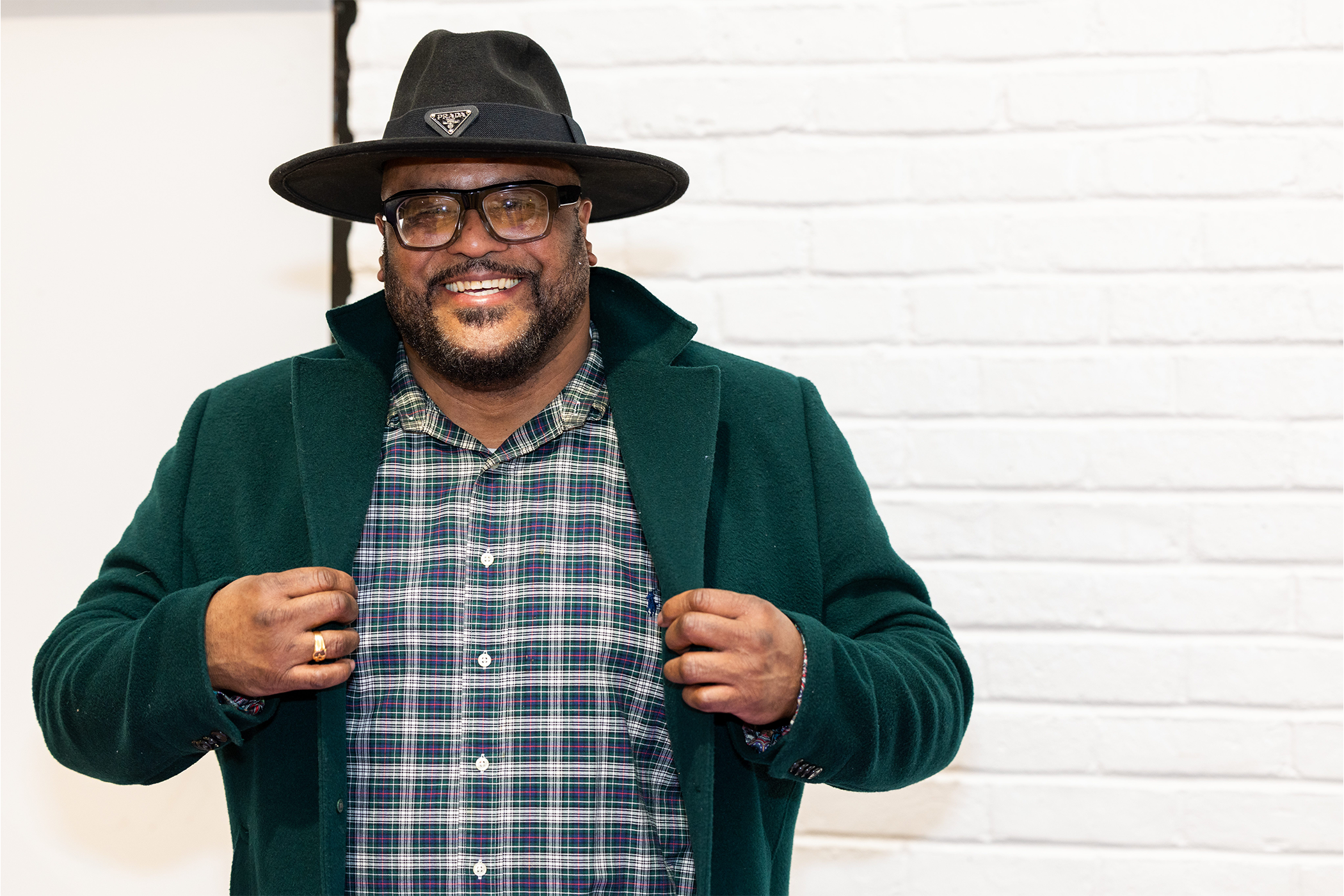 A person wearing a dark green coat over a plaid shirt and a black hat stands in front of a white brick wall.
